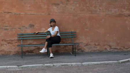 Woman in hat, tshirt, black pants and sneakers sitting on park bench with legs crossed in Bologna...