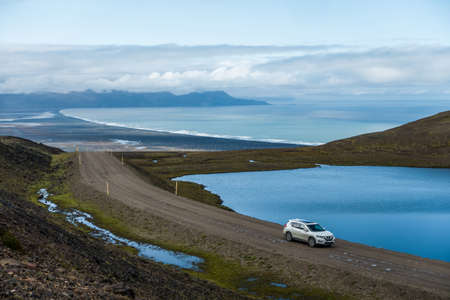 A car passing driving in the road known as Borgarfjardarvegur on the way to Borgarfjordur, passing by a small lake on top of a hill with the ocean in the background. Vatnsskard Eystra, Icelandのeditorial素材