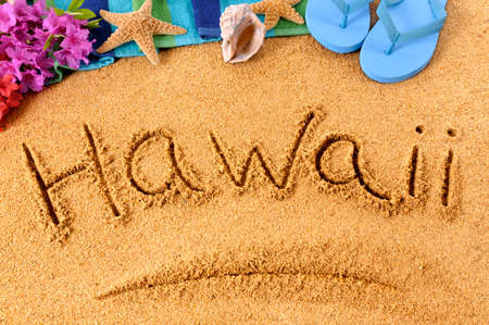 The word Hawaii written on a sandy beach, with flowers, beach towel, starfish and flip flops.の写真素材