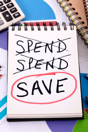 The word Save circled in red below a list of spending written on a notepad surrounded by pencils, graphs, books and calculator.の写真素材