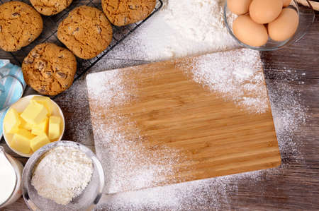 Freshly made chocolate chip cookies on a dark wood table with chopping board and ingredients.の写真素材