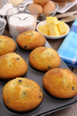 Baking ingredients on a wood table with freshly baked muffins (focus is on the two muffins at the...