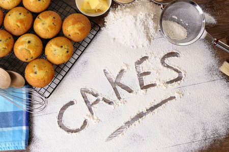 The word Cakes written in flour on a dark wood table with freshly baked muffins and ingredients.