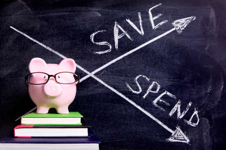 Pink piggy bank with glasses standing on books next to a blackboard with simple spend and save message.  Sharp focus on the piggy bank with blackboard slightly blurred.の写真素材