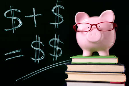 Pink piggy bank with glasses standing on books next to a blackboard with simple money math.  Sharp focus on the piggy bank.の写真素材