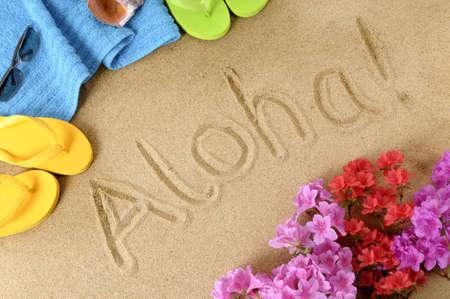 The word Aloha written in soft sand with flip flops, flowers and beach towelの写真素材
