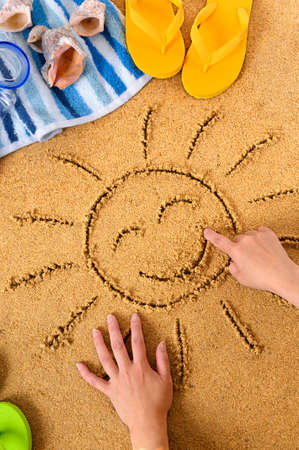 Child drawing a smiley sun in sand with towel, seashells and flip flopsの写真素材