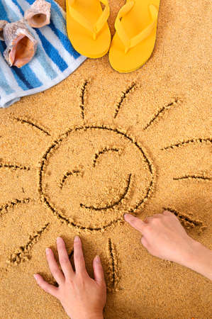Child drawing a smiley sun in sand with towel, seashells and flip flops