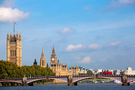 Landscape view of the UK Houses of Parliament looking down the River Thames towards Westminster.  Blue summer sky and Union Jack flag flying in the wind.  Red double decker bus on Lambeth Bridge.  Space for copy.の写真素材