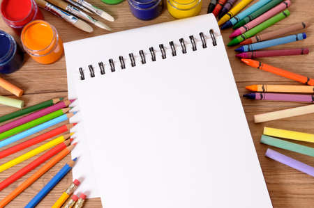 Blank white folded notebook on a school desk with various paints, crayons and pencils.の写真素材