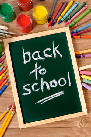 The words Back to School written on a small elementary blackboard with various paints, crayons and pencils on a school desk.の写真素材