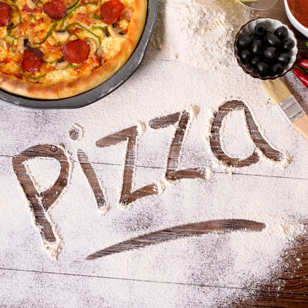 The word Pizza written in flour, with freshly baked pepperoni Pizza surrounded by various ingredients.の写真素材