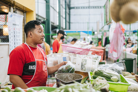 An Indonesian seller who wear red T-shirt and apron is selling groceries in traditional market with bokehのeditorial素材