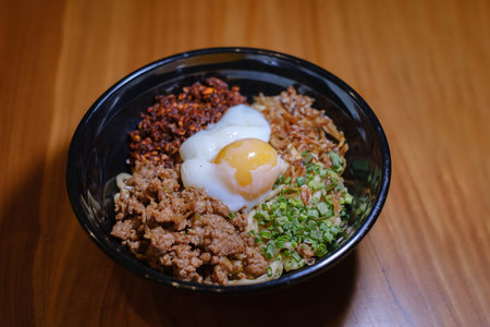 A bowl of Chili Pan Mee containing Half-Cooked Egg, Anchovy Fish, Dried Chili, and Chicken Meat on a wooden tableの写真素材