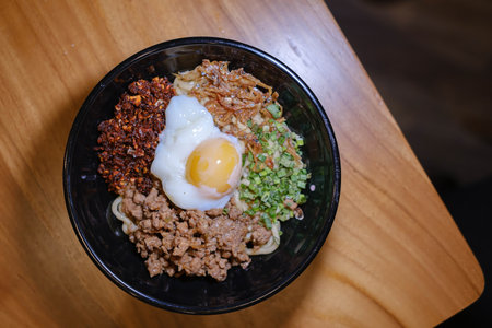 Flat lay or top view shot of a bowl of Chili Pan Mee containing Half-Cooked Egg, Anchovy Fish, Dried Chili, and Chicken Meat on a wooden tableの写真素材