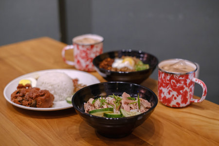 A set of Malaysian cuisine containing Steamed Chicken Rice, Nasi Lemak, Chili Pan Mee, and two mugs of Thai Tea on a wooden table with blurred backgroundの写真素材