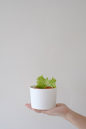 Portrait or vertical shot of a hand holding a white disposable paper bowl with a bunch of fried chickens and a vegetable in it. Isolated white background.の写真素材