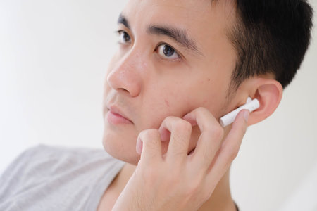 Close-up shot of the partial face of a young Asian man listening to white true wireless stereo earphones. Focus on the eye. Unfocused or shallow depth-of-field. Isolated white background.の写真素材