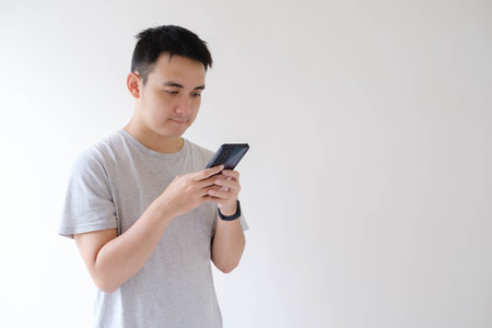 A young Asian man who wears a grey t-shirt and a smartwatch on his wrist is holding and looking at a...