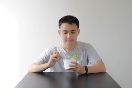A young Asian man wearing a grey T-shirt is eating healthy food on a disposable white paper bowl. Isolated white background.の写真素材