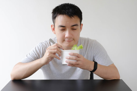 A young Asian man wearing a grey shirt and a smartwatch on his left wrist is smelling a food on disposable paper bowl containing a vegetable. Isolated white background.の写真素材