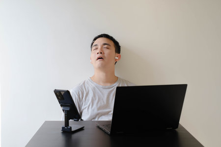 A young Asian man wearing a grey t-shirt and a pair on wireless earphone is falling asleep while working from home. Laptop and smartphone on the black table. Isolated white background.の写真素材