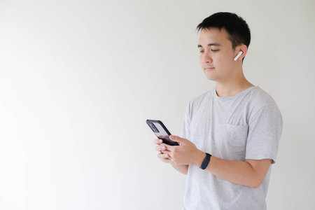 A young Asian man who wears a grey t-shirt, using a pair of wireless earphones and a smartwatch on his left wrist is holding and looking at a smartphone while listening to music on his wireless earphones. Isolated white background.の写真素材