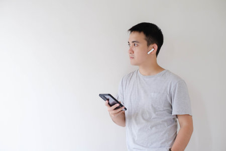 A young Asian man who wears a grey t-shirt, a pair of wireless earphones, and a smartwatch is holding a smartphone and looking at the left side. Isolated white background.の写真素材
