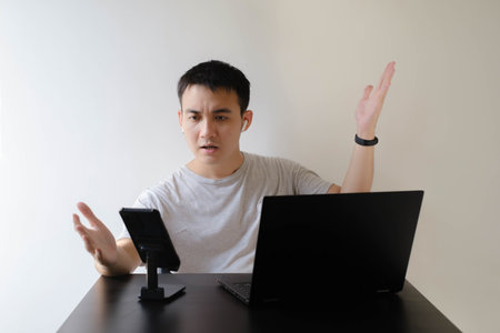A young Asian man wearing a gray t-shirt, a smartwatch on his left wrist, and a pair of wireless earphones is angry at an online meeting. Laptop and phone on wooden table. Isolated white background.の写真素材