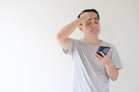 Medium and soft-focused shot of a young Asian man who wears a grey t-shirt and is facepalm while holding while looking at a smartphone. Shallow depth-of-field. Isolated white background.の写真素材