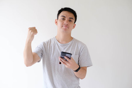 A young Asian man wearing a grey T-shirt and a smartwatch on his left wrist is happy with a flat expression while holding a smartphone. Isolated white background.の写真素材