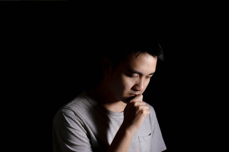 Low key shot of a young Asian man is overthinking with his head down. Isolated black background. Dramatic.の写真素材