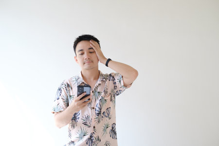A young Asian man wearing a pink shirt with tropical patterns and a smartwatch on his left wrist feels brainstormed when looking at the smartphone. Isolated white background.の写真素材