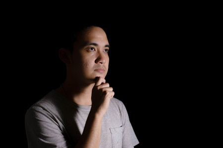 Low key shot of a young Asian man wearing a grey T-shirt is in a thinking pose. Isolated black background. Dramatic.の写真素材