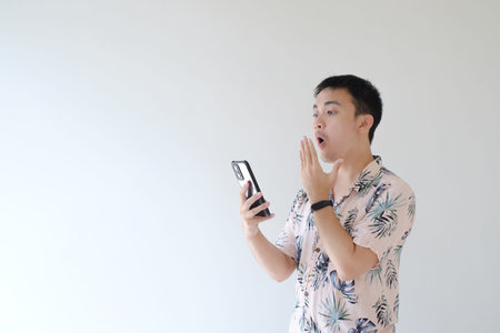 A young Asian man wearing a pink shirt with tropical patterns is holding a smartphone with a shocked expression. Isolate white background.の写真素材