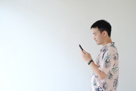 A young Asian man wearing a pink shirt with tropical patterns and a smartwatch on his left wrist is using his smartphone facing leftの写真素材