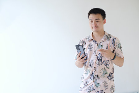 A young Asian man wearing a pink shirt with tropical patterns and a smartwatch on his left wrist is holding a smartphone and pointing at it. Isolated white background.の写真素材