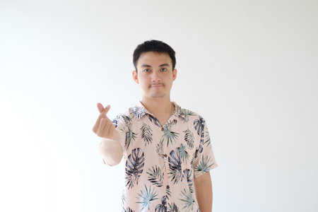 A young Asian man wearing a pink shirt with tropical patterns is posing as "Saranghae" usually known as finger heart. Focused on the man. Unfocused hand. Isolated white background.の写真素材