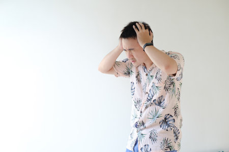 A young Asian man wearing a pink colored shirt with tropical patterns and a smartwatch on his left wrist is feeling dizzy. Isolated white background.の写真素材