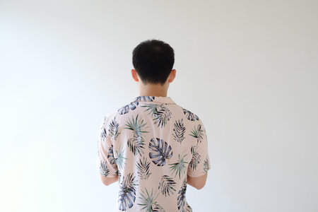 A young Asian man wearing a pink shirt with tropical patterns is standing with his back on the...