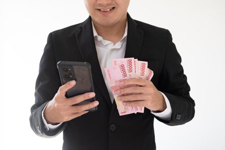 Close-up shot of a partial face of a young Asian businessman is holding a smartphone and a bunch of Indonesian money with happy expression. Isolated white background. Suitable for advertisement.の写真素材