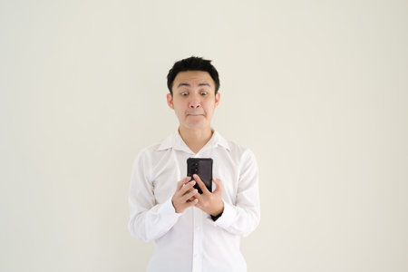 Young Asian man wearing a white shirt and a watch holding a smartphone with shocked face expression. Isolated white background.の写真素材