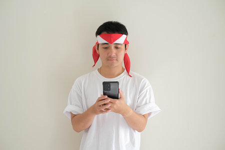 A young Asian man wearing a white T-shirt and Kemerdekaan Indonesia costume is looking at his smartphone. Indonesia Independence Day. Isolated white background.の写真素材