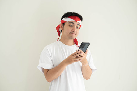 A young Asian man wearing a white T-shirt and Kemerdekaan Indonesia costume is looking at his smartphone with flat expression. Indonesia Independence Day. Isolated white background.の写真素材