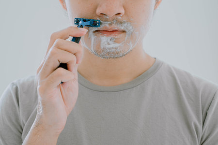 Selective focus and close-up shot of a man wearing sage green casual T-Shirt is shaving his mustache using shaver and cream with flat expression on isolated white backgroundの写真素材