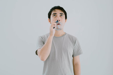 Selective focus shot of young Asian man wearing sage green casual T-Shirt is shaving his mustache using shaver and cream with flat expression while looking up on isolated white background.の写真素材