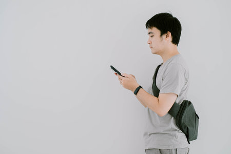 Selective focus shot of young Asian man wearing sage green T-Shirt, a smartwatch, and minimalist sling bag is using smartphone while facing on the left side. Isolated white background.の写真素材