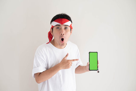 A young Asian man wearing a white T-shirt and Kemerdekaan Indonesia outfit is pointing to the smartphone with green screen. Indonesian Independence Day. Suitable for advertisement.の写真素材