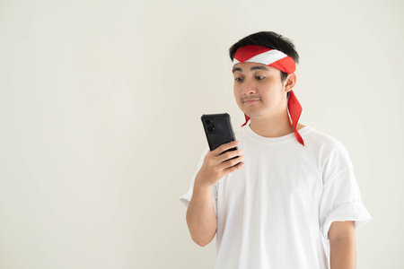 A young Asian man wearing a white T-shirt and Kemerdekaan Indonesia costume is looking at his smartphone with flat expression. Indonesia Independence Day. Isolated white background.の写真素材