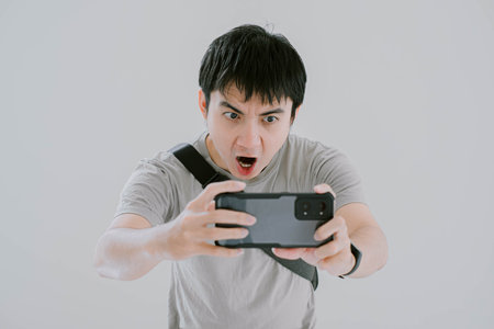 Selective focus shot of young Asian man wearing sage green casual T-Shirt, smartwatch, and minimalist sling bag is capture photo using smartphone with excited expression on isolated white backgroundの写真素材